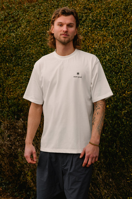 Man wearing a white t-shirt with a logo against a green hedge background