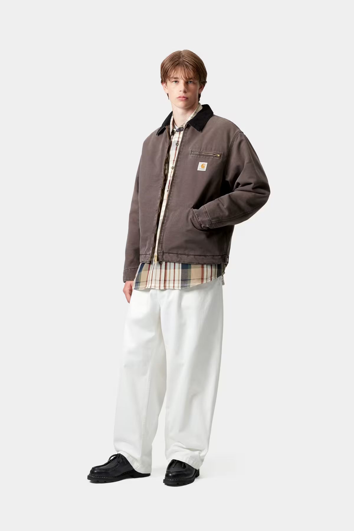 Person wearing a brown jacket, plaid shirt, and white pants on a white background