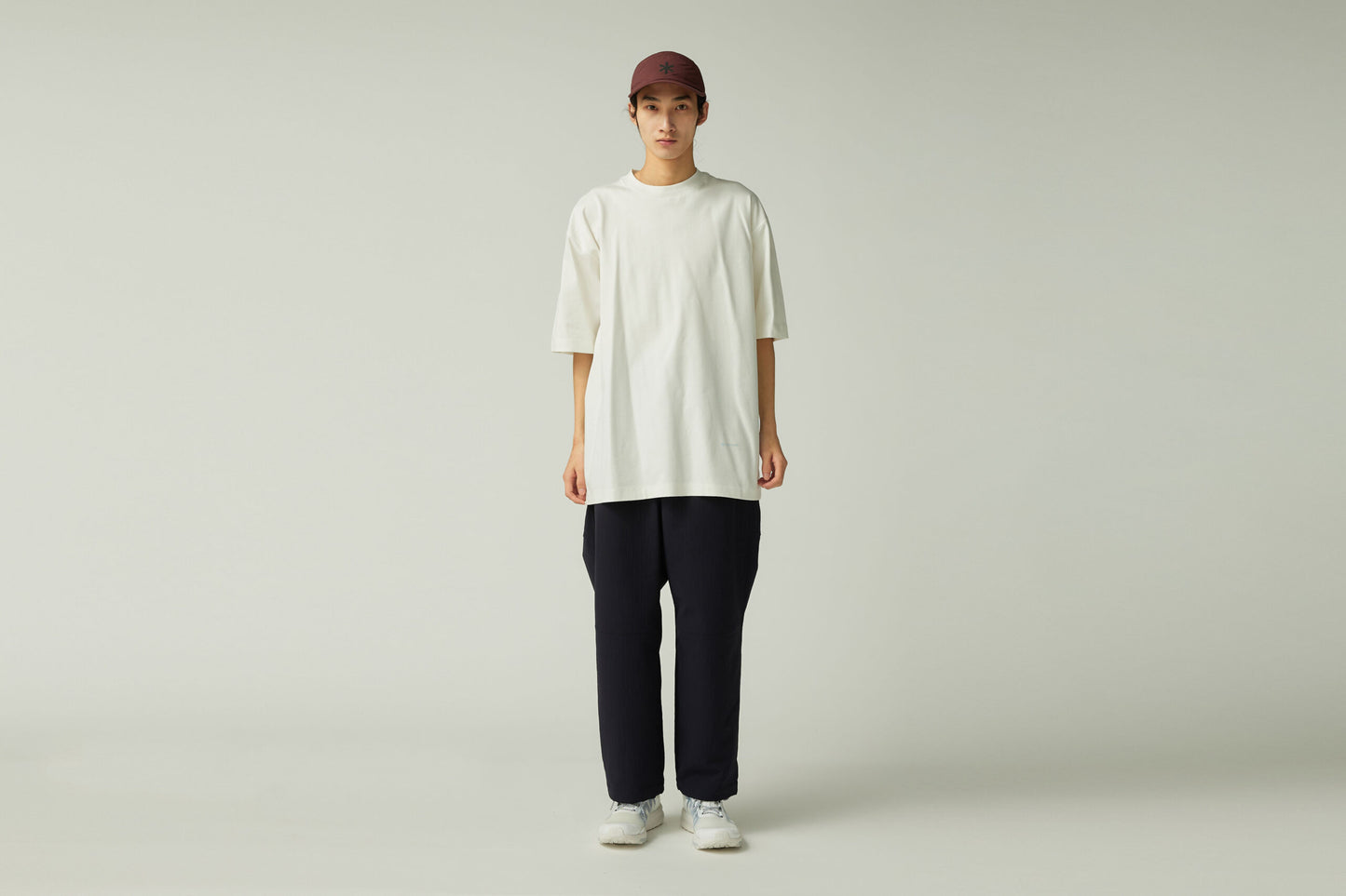 Person wearing a white oversized t-shirt, black pants, and a maroon cap on a plain background