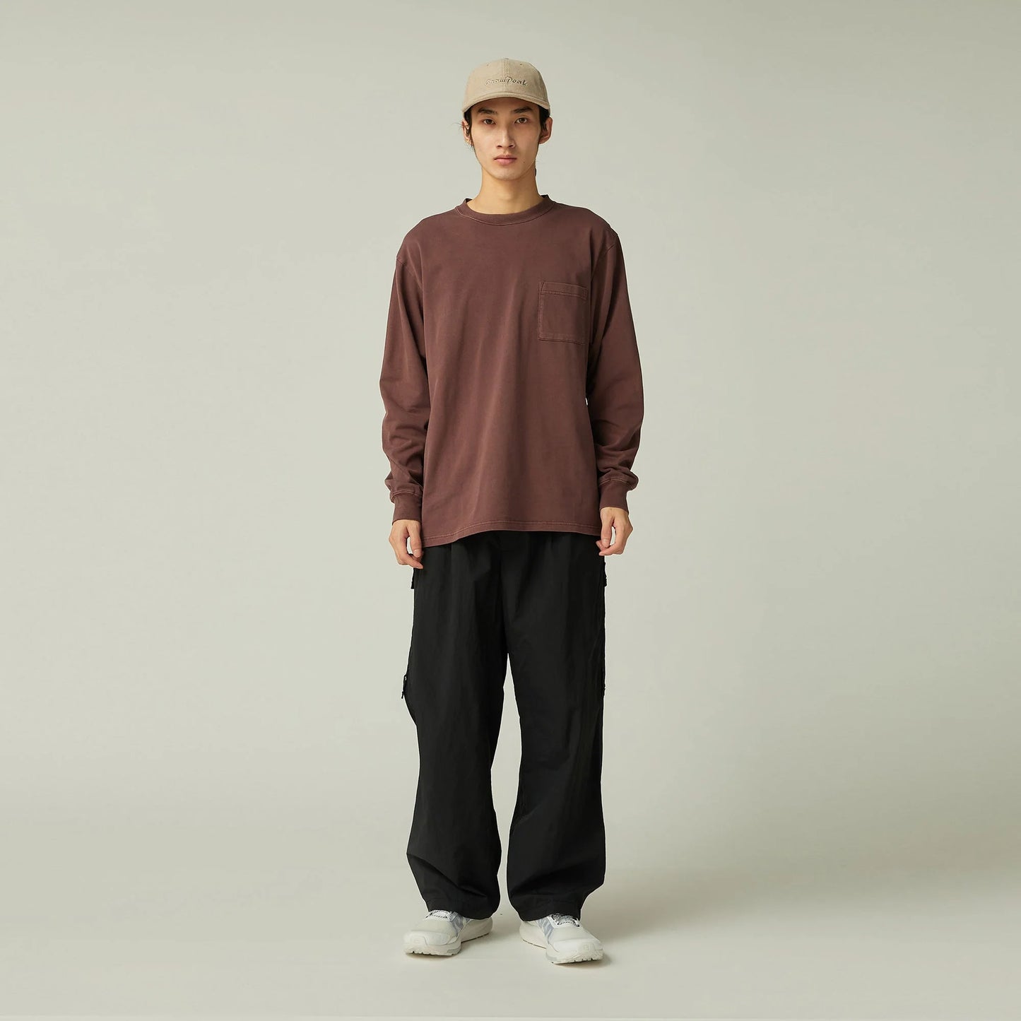 Person wearing a brown long-sleeve shirt, black pants, and a beige cap on a plain background